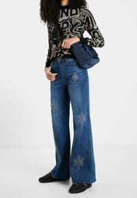 Jean en denim bleu à jambes larges avec des motifs de fleurs brodées. Associé à un pull noir à motifs et un sac à bandoulière bleu marine. Chaussures noires.