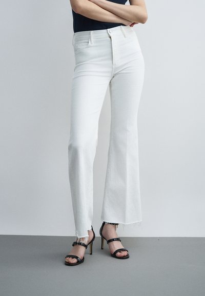 Person mit hoch taillierter weißer Jeans mit rohem Saum und schwarzen, riemchenbesetzten High-Heel-Sandalen, die mit verschränkten Armen vor einem schlichten Hintergrund steht.