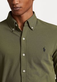 Tmavě zelená košilová halenka vyrobená z texturované tkaniny, s klasickým límcem a malým černým logem na levé hrudi, s bílými knoflíky.