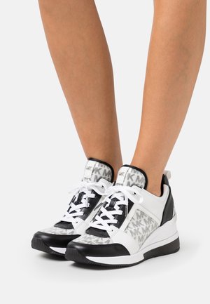 Personne portant des baskets de marque noires, blanches et grises avec des lacets, debout sur un fond clair uni.