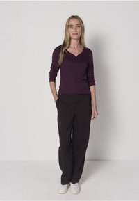 Long-sleeve, deep purple top with a v-neck, paired with loose-fitting black trousers. Model wears white sneakers, with smooth textures shown.