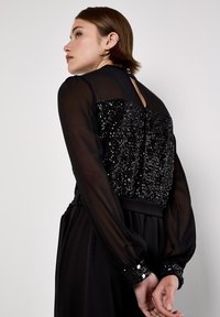 Schwarzes Kleid mit paillettenbesetztem Oberteil, transparenten langen Ärmeln und einem Dekolleté-Rücken. Plissierter Rock und Knopfdetails an den Manschetten.