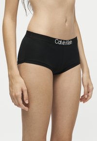 Γυναίκα που φοράει μαύρα boyshort εσώρουχα Calvin Klein, εμφανίζεται από τη μέση έως τη μέση του μηρού, στέκεται μπροστά σε ένα απλό ανοιχτό φόντο.