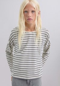 Joven con cabello largo y rubio que lleva una camisa blanca de manga larga con rayas horizontales negras y pantalones grises, de pie contra un fondo liso.