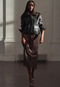 Frau, die eine schwarze Lederjacke über einem schimmernden bronzefarbenen Spitzenkleid und goldenen Sandalen trägt, steht in einem sanft beleuchteten Raum mit klassischen Details.