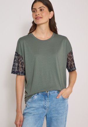 Kvinde iført en løs grøn t-shirt med sorte leopardprint netærmer og lyseblå cowboybukser, stående med den ene hånd i lommen.