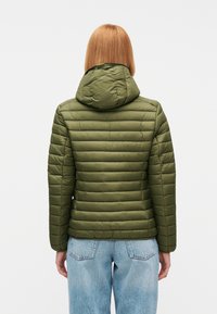 Άτομο με ίσια ανοιχτό καστανά μαλλιά που φοράει μια λαδοπράσινη καπιτονέ ζακέτα με κουκούλα και ανοιχτό μπλε τζιν παντελόνι, βλέπουμε από πίσω.