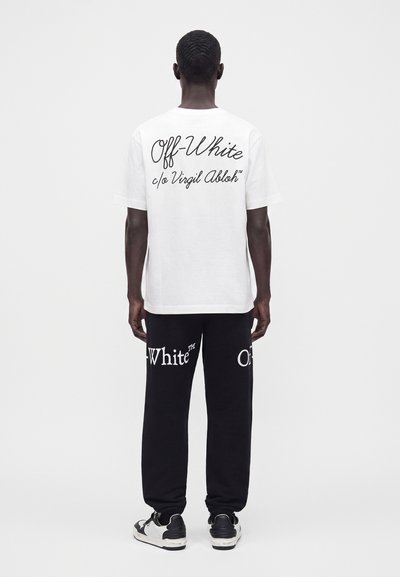 Homme debout de dos, portant un t-shirt blanc Off-White et un pantalon noir de la marque Off-White avec des baskets noires et blanches.