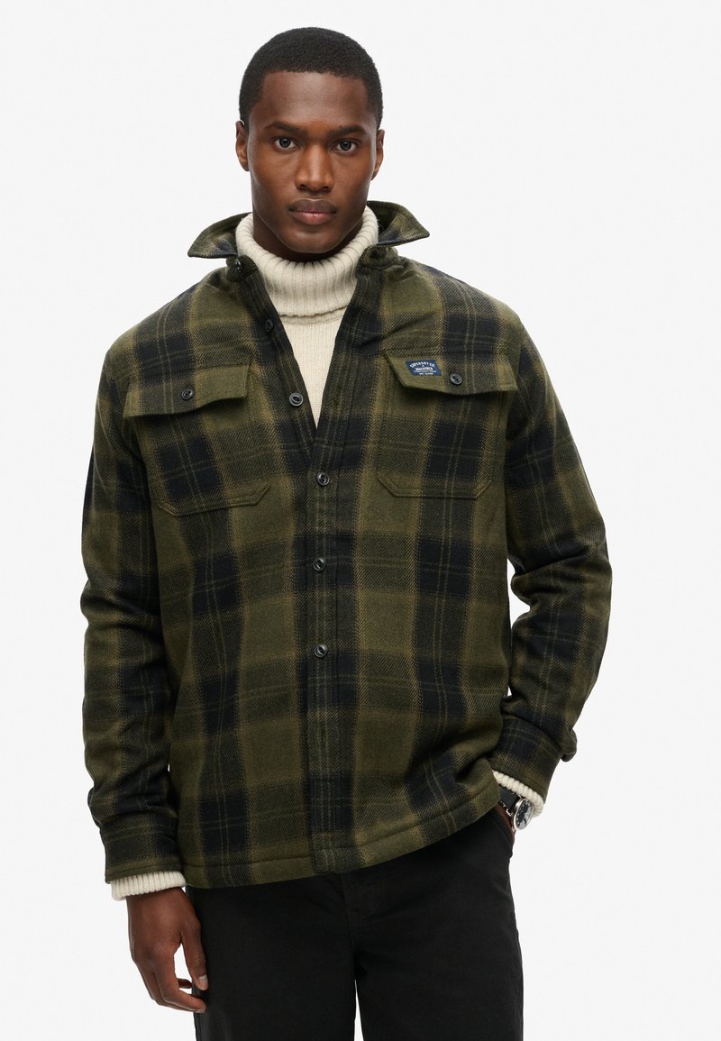 Camisa de franela en cuadro verde y negro, con cierre de botones al frente, dos bolsillos en el pecho y puños acanalados, usada sobre un cuello de tortuga color crema.