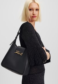 Black leather handbag with a structured design, gold logo accent on the flap, and a single adjustable strap. Worn against a sparkly black outfit.