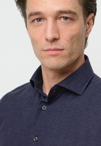 Camisa azul oscuro de botones con una tela texturizada, que presenta un cuello extendido y dos botones negros visibles en el frente.