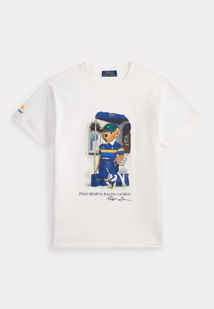 Kvinde iført solbriller, hvid kabelstrikket lynlås-hættetrøje, hvid T-shirt med bjørngrafik, navy plisseret nederdel, hvide sokker og hvide sneakers.
