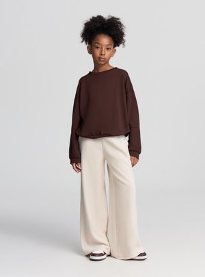 Felpa marrone oversize con maniche larghe, abbinata a pantaloni crema a gamba larga. Scarpe da ginnastica bianche e marroni completano l'outfit.