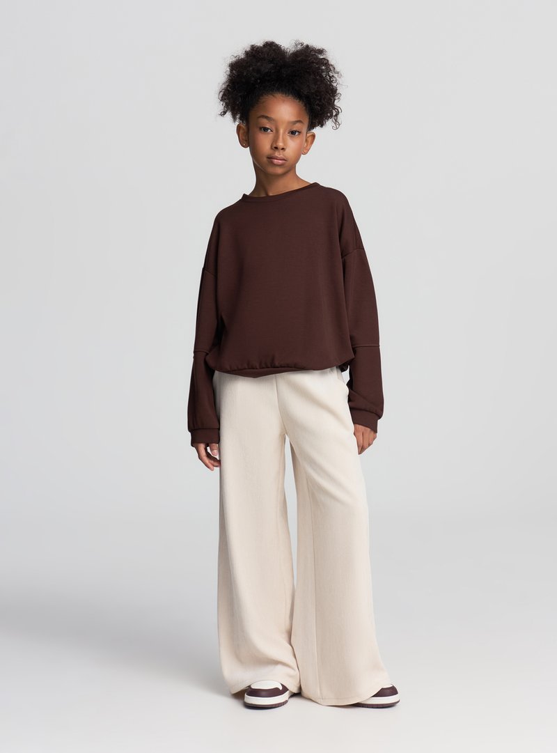 Felpa marrone oversize con maniche larghe, abbinata a pantaloni crema a gamba larga. Scarpe da ginnastica bianche e marroni completano l'outfit.