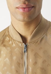 Veste bomber beige à texture lisse, dotée d'une fermeture éclair à l'avant, d'un col côtelé et d'un motif tonal subtil. Détails des fermetures éclair en argent visibles.