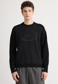 Maglione nero con un logo testurizzato al centro, scollatura rotonda e maniche lunghe. Tessuto liscio con un design aderente. Chi lo indossa ha i capelli corti e ricci.