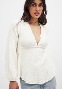 Witte geëtaleerde blouse met een diepe V-hals, opbollende lange mouwen en een gebogen zoom. De stof heeft een gegolfde afwerking, wat de visuele interesse vergroot.