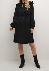 Svart satin klänning med långa ärmar, volangdetaljer på axlarna och dragsko vid halsringningen. Har en avslappnad passform och knä-lång nederkant.