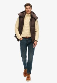Braune Steppweste über einem cremefarbenen, strukturierten Pullover, blaue Jeans und braune Schnürstiefel; lässiges Outfit mit einem geschichteten Look.