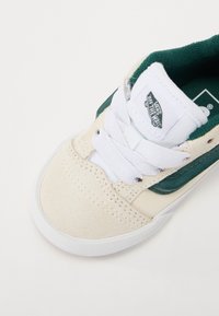 Beżowe zamszowe sneakersy z zielonymi skórzanymi akcentami, białymi sznurówkami i wyściełanym językiem. Zawierają naszywkę z logotypem na języku.