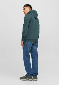 Sudadera de color verde oscuro con un acabado texturizado, bolsillo canguro en la parte frontal y capucha con cordón ajustable. Llevada con jeans de mezclilla azules y zapatillas grises.