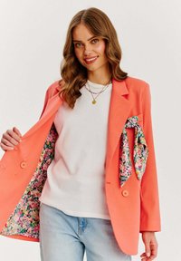 Korálový blazer s podšívkou s kvetinovým vzorom, kombinovaný s bielou košeľou a svetlo modrými džínami. Farebný šál dopĺňa blazer.