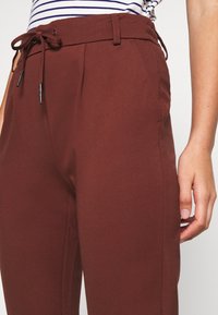 Burgundin väriset housut, joissa on naru vyötäröllä, laskokset ja sivutaskut; valmistettu sileästä kankaasta, jossa on muotoiltu istuvuus ja hienovarainen tekstuuri.