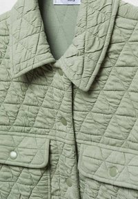 Veste matelassée vert clair avec grand col, boutons-pression à l'avant et poches à rabat, montrant un motif de quilting en losanges texturé.