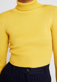 Maglione giallo a collo alto in tessuto morbido dal design aderente. Presenta maniche lunghe e colletto alto, abbinato a pantaloni scuri a righe sottili.