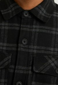 Black and gray plaid shirt with a collar, featuring four buttons and two chest pockets, made of soft, textured fabric.