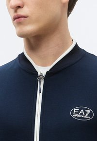 Giacca con zip blu navy con dettagli bianchi, colletto a coste e logo EA7 Emporio Armani. Presenta un tessuto strutturato e una cerniera argentata.