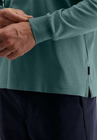 Mann trägt ein türkisfarbenes Langarmshirt und dunkle Hosen, während er seinen Ärmel mit gefalteten Händen anpasst.