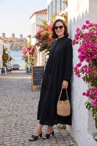 Donna con un lungo vestito nero e occhiali da sole in piedi contro un muro bianco con fiori rosa su una strada lastricata vicino al mare.