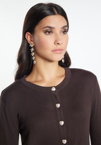 Bruine cardigan met een ronde halslijn, voorzien van hartvormige knopen aan de voorkant. G gedragen met hangende bloemenoorbellen. Gladde textuur.