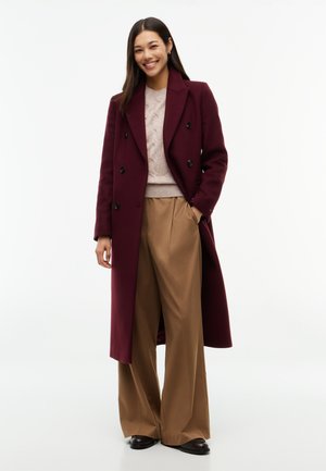 Femme portant un long manteau bordeaux, un pull beige, un pantalon large marron et des chaussures noires, se tenant debout et souriante sur fond blanc.