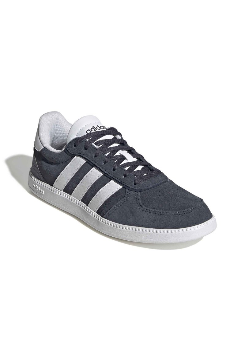 Zapatilla de ante gris oscuro con rayas de cuero blanco, punta redonda, parte delantera con cordones, cuello acolchado y suela de goma. Logo de la marca en el lateral.
