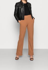 Svart läderjacka med tryckknappar, åtsittande svart topp och bruna högmidjade vida byxor. Svarta klacksandaler. Släta texturer och rena linjer.