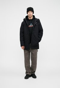 Ung mann som står iført svart lue, svart hettegenser, grafisk hettegenser, grå bukser med knepatsch, og svarte joggesko foran hvit bakgrunn.