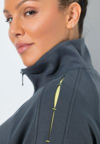 Mujer con chaqueta gris oscuro con cremallera y cuello alto, con detalles en recorte amarillo en el hombro, mirando de lado.