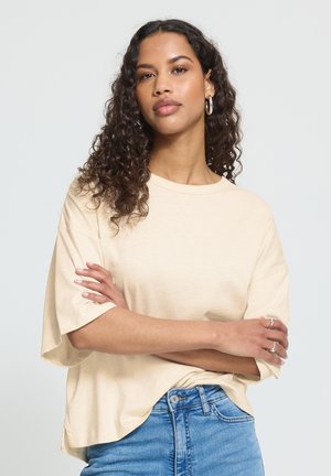 Kvinde med krøllet hår iført en beige oversized t-shirt og blå jeans, stående med armene krydsede mod en ensfarvet baggrund.