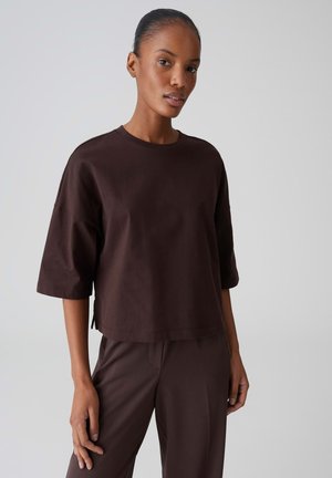 Woman wearing a loose dark brown short-sleeve shirt and matching pants, standing against a plain light gray background.