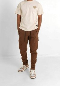 Man i beige t-shirt med hjärtpatch, bruna cargobyxor med fickor och beige och vita sneakers står med händerna i fickorna.