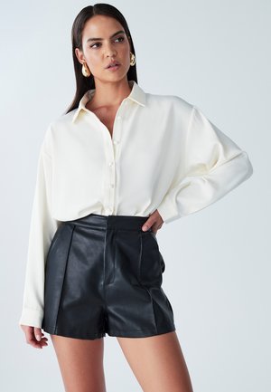 Blusa de crema con diseño de botones y ajuste relajado, combinada con pantalones cortos de cuero negros de talle alto con pliegues y bolsillos laterales.