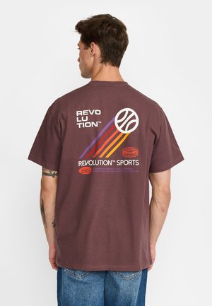 Man draagt een bruin T-shirt met de tekst "Revolution Sports" en een kleurrijke basketbalafbeelding op de achterkant, gecombineerd met een blauwe spijkerbroek.