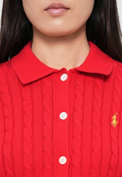 Rdeča pletena polo majica s teksturo kabla, ovratnikom, tremi belimi gumbi in majhnim zlatim logotipom.
