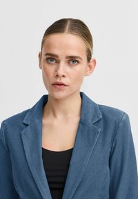 Ljusblå kavaj med slagkrage och slät textur, bärs över en svart topp. Passformen är strukturerad, vilket framhäver axlarna.