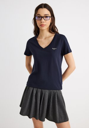 Woman wearing navy blue V-neck T-shirt and gray pinstriped pleated skirt, standing with hands behind back, neutral background.