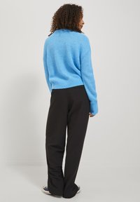 Svetlo modri pleteni pulover z ohlapnim krojem, rebrasto teksturo in spušenimi rameni, v kombinaciji z črnimi širokimi hlačami in priložnostnimi čevlji.