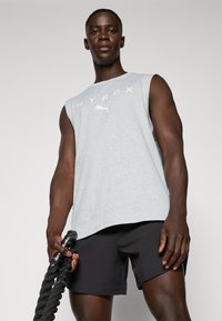 Lys grå ermeløs skjorte med "HYROX" i hvit tekst; kombinert med svarte shorts. Modellen holder et tykt, teksturert svart treningstau.