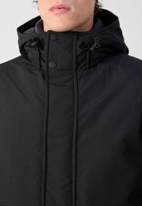 Gros plan d'une personne portant une veste à capuche noire avec des boutons et un cordon au niveau du col.